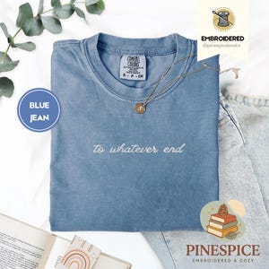May include: A blue jean coloured t-shirt with the phrase "to whatever end" embroidered in white. A gold necklace with a pendant rests on the shirt. The shirt is folded on top of a pair of jeans.