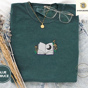 May include: A blue spruce t-shirt with an embroidered design of an open book, a crescent moon, and mushrooms. The shirt has a crew neck and is adorned with a gold necklace and clear glasses. The text "Embroidered" and "Blue Spruce" are visible.