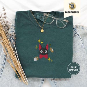 Może przedstawiać: Bluza w kolorze niebieskiego świerku z haftowanym czarnym kotem, czerwoną muszką i złotymi gwiazdami. Bluza jest połączona ze złotym naszyjnikiem i okularami w przezroczystych oprawkach. Słowo "EMBROIDERED" jest wydrukowane w prawym górnym rogu.
