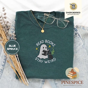 Op de afbeelding: Een blauwspar t-shirt met een geborduurd ontwerp met een spook dat een boek leest naast een zwarte kat, met de tekst "READ BOOKS STAY WEIRD". Een gouden ketting en een doorzichtige bril liggen op de trui.
