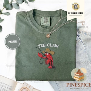 May include: Moss green t-shirt with the embroidered phrase "Yee-Claw" and a cartoon lobster wearing a cowboy hat. The shirt is a Comfort Colors brand, size S/P/CH. A gold necklace with a pendant is draped on the shirt.