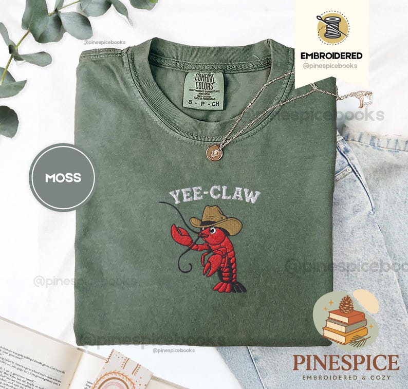 May include: Moss green t-shirt with the embroidered phrase "Yee-Claw" and a cartoon lobster wearing a cowboy hat. The shirt is a Comfort Colors brand, size S/P/CH. A gold necklace with a pendant is draped on the shirt.