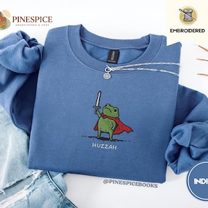 May include: Indigo blue sweatshirt with an embroidered design of a green frog holding a sword and wearing a red cape. The word "Huzzah" is embroidered below the frog. The sweatshirt has a crew neck and long sleeves.
