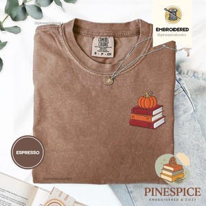 以下が含まれることがあります： エスプレッソ色のTシャツ。刺繍されたカボチャと本のデザイン。クルーネックと半袖。テキスト「Comfort Colors」が見えます。ペンダント付きのシルバーネックレスがTシャツの上にあります。