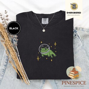 Könnte beinhalten: Schwarzes Comfort Colors T-Shirt mit einer gestickten grünen Frosch-Illustration, der einen Weltraumhelm trägt, umgeben von gelben Sternen. Der Schriftzug "BLACK" befindet sich in einem schwarzen Kreis. Der Text "EMBROIDERED @pinespicebooks" ist sichtbar.