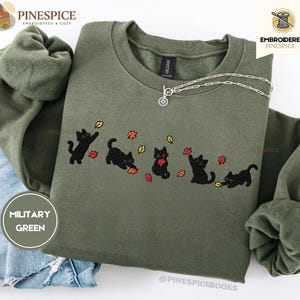 May include: A military green crewneck sweatshirt featuring an embroidered design of five black cats playing with scattered red, orange, and yellow autumn leaves. The sweatshirt has long sleeves with ribbed cuffs and a ribbed neckline.