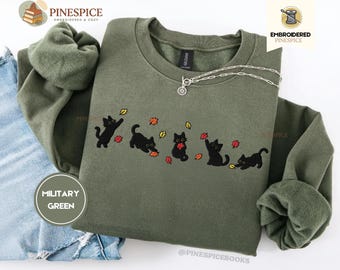 Puede incluir: Una sudadera de cuello redondo color verde militar con un diseño bordado de cinco gatos negros jugando con hojas de otoño rojas, naranjas y amarillas. La sudadera tiene cuello y puños acanalados, y un collar de tono plateado con cierre.