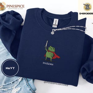 May include: Navy blue sweatshirt with an embroidered design of a green frog holding a sword and wearing a red cape. The word "Huzzah" is embroidered below the frog. The sweatshirt has a crew neck and long sleeves.