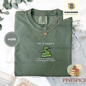 Puede incluir: Una camiseta color verde musgo con un bordado de una serpiente verde que lleva una bufanda a rayas amarillas y verdes. El texto dice I'M A SNAKE y I'M A SLITHERING LITTLE SNAKEY SNAKE. La camiseta tiene una etiqueta Comfort Colors en el cuello.