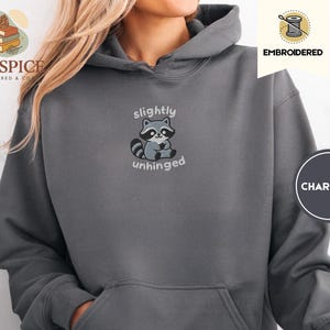 Pu&ograve; includere: Felpa con cappuccio grigio antracite con un design ricamato di procione e le parole "slightly unhinged". La felpa ha un cappuccio con coulisse e una tasca frontale. Il marchio "Pinespice" &egrave; visibile in alto a sinistra.