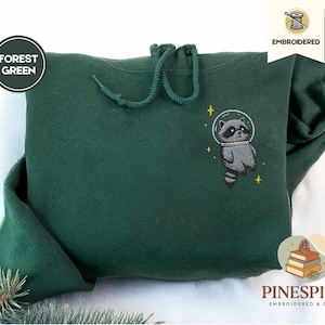 以下が含まれることがあります： 宇宙ヘルメットをかぶったアライグマの刺繍が施された、フォレストグリーンのパーカー。パーカーにはドローストリングフードとカンガルーポケットが付いています。「Forest Green」の文字が左側の円の中にあります。