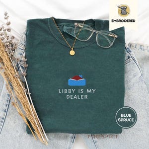以下が含まれることがあります： 「LIBBY IS MY DEALER」の刺繍と家のデザインが施された、ブルー・スプルース色のTシャツ。ゴールドのネックレス、クリアグラス、デニムジーンズとコーディネートされています。右上には「EMBROIDERED」の文字があります。