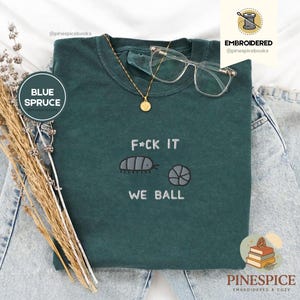May include: Blue Spruce t-shirt with the embroidered phrase "F*CK IT WE BALL" and a pill bug and ball graphic. A gold necklace with a pendant and clear glasses rest on the shirt. The shirt is on a pair of blue jeans.