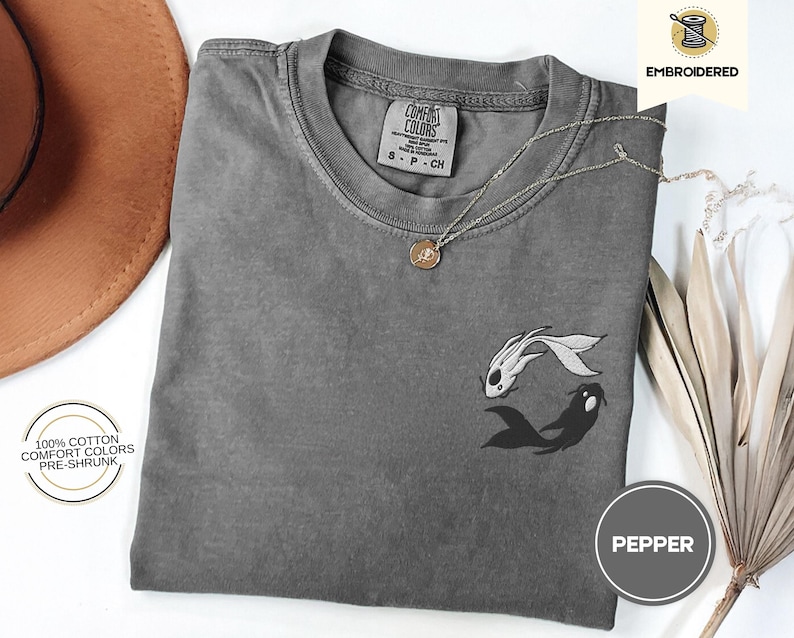 May include: A gray t-shirt with a black and white embroidered design of two fish. The text "COMFORT COLORS" and "8-P-CH" is visible on the tag. The text "100% COTTON COMFORT COLORS PRE-SHRUNK" and "PEPPER" are also visible.