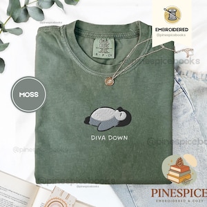 May include: A moss green t-shirt with an embroidered design of a sleeping creature and the words "DIVA DOWN". The shirt has a crew neck and short sleeves. A gold necklace is draped on the shirt.