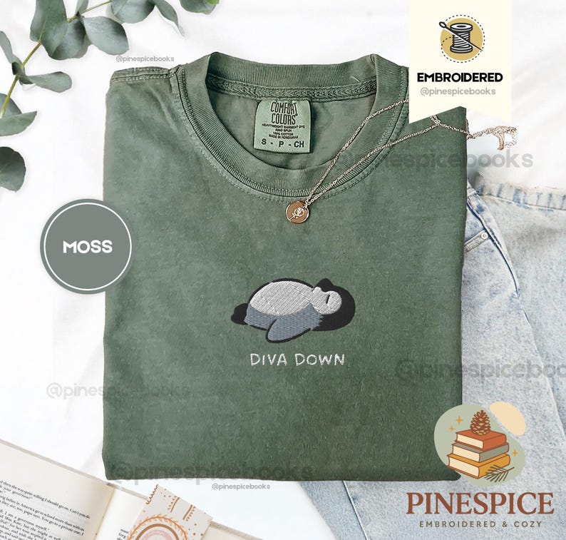May include: A moss green t-shirt with an embroidered design of a sleeping creature and the words "DIVA DOWN". The shirt has a crew neck and short sleeves. A gold necklace is draped on the shirt.