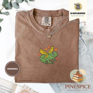 May include: Espresso-colored t-shirt with a crew neck and short sleeves. The shirt features an embroidered green frog with yellow wings and red star accents. The shirt has the Comfort Colors brand label and a small, round pendant necklace.