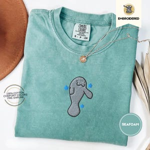 May include: Seafoam green Comfort Colors t-shirt with an embroidered manatee design and blue star accents. The shirt is labelled "Embroidered" and "Seafoam" with "100% Cotton Comfort Colors Pre-Shrunk" text. A silver necklace with a pendant is also visible.