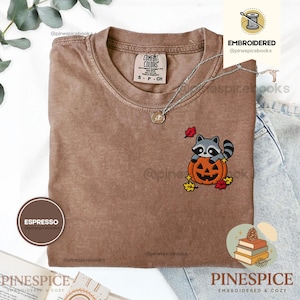 Pode incluir: Uma t-shirt cor de café expresso com um design bordado de um guaxinim sentado dentro de uma abóbora laranja esculpida. O bordado inclui pequenas folhas de outono amarelas e vermelhas ao redor da abóbora. A t-shirt tem gola redonda.