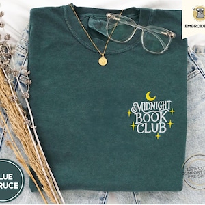 Op de afbeelding: Een donkergroen T-shirt met de tekst "Midnight Book Club" geborduurd in geel op de voorkant. De tekst wordt omringd door kleine gele sterren.