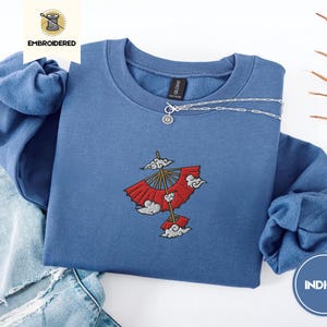 May include: Indigo blue sweatshirt featuring an embroidered design of a red and white umbrella with clouds. The sweatshirt has a crew neck and long sleeves.
