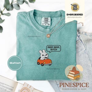 May include: Seafoam green t-shirt with an embroidered design of a gray bunny in an orange car, with the text "BEEP BEEP, BITCH" in a speech bubble. The shirt also has the text "PINESPICE EMBROIDERED & COZY".