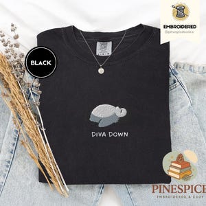 May include: Black crew neck sweatshirt with an embroidered turtle design and the text "DIVA DOWN". The sweatshirt has a round black label that says "BLACK". A silver necklace with a pendant is visible.
