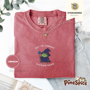 Può includere: Una t-shirt a maniche corte color cremisi con un ricamo di una rana verde che indossa un cappello e una tunica da mago blu scuro, tenendo un bastone con una sfera blu. Il testo sopra e sotto la rana recita, Off to commit shenanigans.