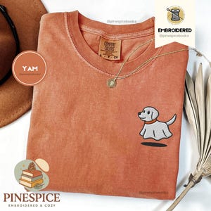 May include: A terracotta-colored t-shirt with a small embroidered cartoon dog. The shirt has a crew neck and a small tag. A gold necklace with a pendant is draped on the shirt. The brand name "PINESPICE" is printed at the bottom.