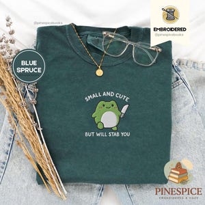 Puede incluir: Una camiseta azul abeto con un diseño de rana bordada sosteniendo un cuchillo. El texto "SMALL AND CUTE BUT WILL STAB YOU" está debajo de la rana. Un collar dorado y gafas transparentes están sobre la camiseta.