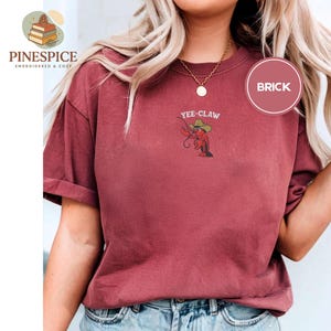 May include: Brick-colored t-shirt with rolled sleeves. Features an embroidered design of a cartoon crawfish wearing a cowboy hat, with the text "Yee-Claw" above it. The shirt also has a small logo on the upper left side.