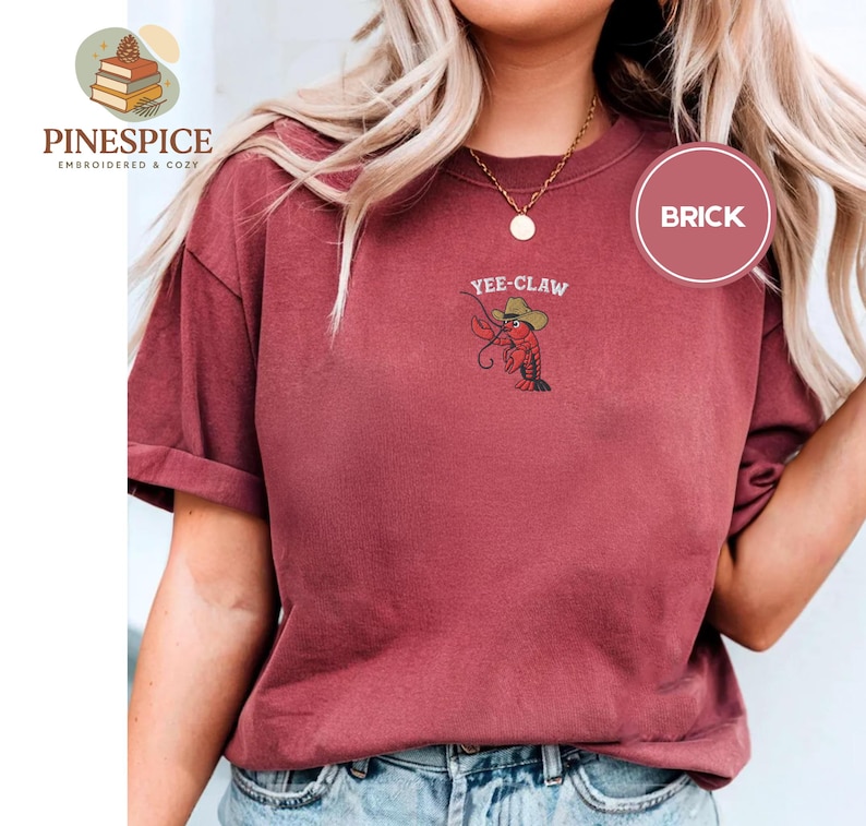 May include: Brick-colored t-shirt with rolled sleeves. Features an embroidered design of a cartoon crawfish wearing a cowboy hat, with the text "Yee-Claw" above it. The shirt also has a small logo on the upper left side.