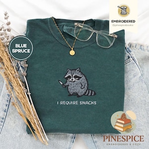 Puede incluir: Camiseta azul abeto con un mapache bordado sosteniendo un cuchillo. El texto "I REQUIRE SNACKS" está debajo. Un collar dorado y gafas transparentes descansan sobre la camiseta. Colección Pinespice.