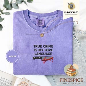 Pode incluir: Camiseta violeta Comfort Colors com a frase "TRUE CRIME IS MY LOVE LANGUAGE" e um gráfico de uma faca com sangue. Um colar dourado está colocado na camiseta. A camiseta é roxa.