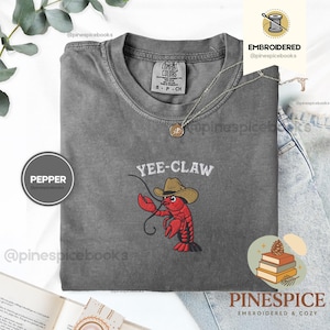 May include: A gray Comfort Colors t-shirt with the embroidered phrase "YEE-CLAW" above a red lobster wearing a cowboy hat. A silver necklace with a pendant is on the shirt. The shirt is size S-P-CH.