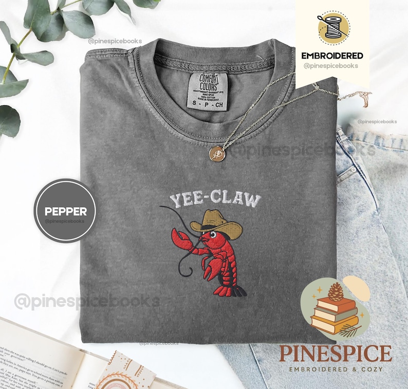 May include: A gray Comfort Colors t-shirt with the embroidered phrase "YEE-CLAW" above a red lobster wearing a cowboy hat. A silver necklace with a pendant is on the shirt. The shirt is size S-P-CH.