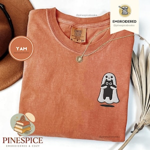 Puede incluir: Una camiseta de color naranja quemado con cuello redondo y mangas cortas. La camiseta presenta un diseño bordado de un gato negro con un disfraz de fantasma blanco. Un collar dorado con un colgante está drapeado sobre la camiseta. La marca "PINESPICE" está impresa en la parte inferior.