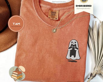 May include: A burnt orange t-shirt with a crew neck and short sleeves. The shirt features an embroidered design of a black cat in a white ghost costume. A gold necklace with a pendant is draped on the shirt. The brand name "PINESPICE" is printed at the bottom.