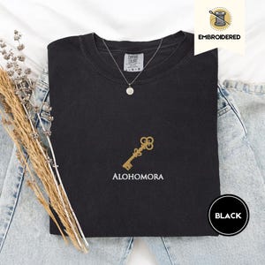 May include: Black crew neck t-shirt with a gold key design and the word "ALOHOMORA" embroidered below it. The shirt is labeled "Comfort Colors" and has a small, round pendant necklace. The word "EMBROIDERED" is printed on the top right.
