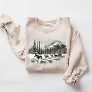 May include: A beige sweatshirt with a watercolor illustration of a snowy mountain scene with a team of huskies pulling a sled. The illustration is in shades of blue, green, and brown.