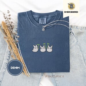 May include: A denim blue t-shirt with an embroidered design of three white cartoon characters with green leaves. The shirt has a crew neck and short sleeves. A silver necklace with a pendant is on the shirt. The word "EMBROIDERED" is printed on the image.
