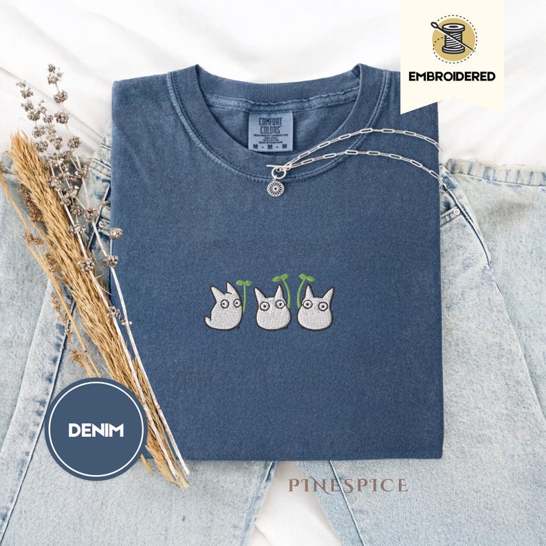 May include: A denim blue t-shirt with an embroidered design of three white cartoon characters with green leaves. The shirt has a crew neck and short sleeves. A silver necklace with a pendant is on the shirt. The word "EMBROIDERED" is printed on the image.