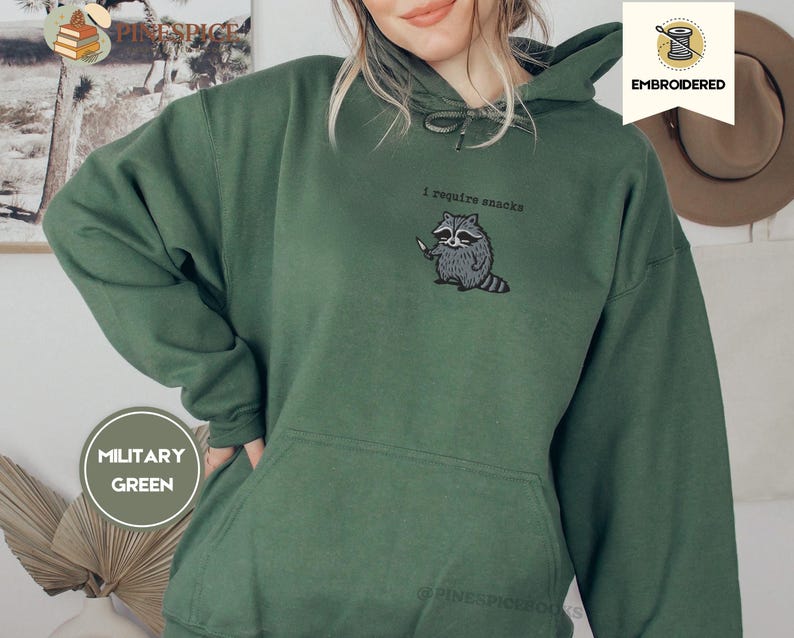 May include: Military green hoodie with a kangaroo pocket and drawstring hood. The embroidered design features a raccoon holding a knife with the text "I require snacks". The hoodie is a comfortable, casual style.