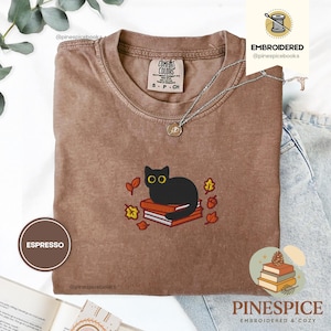 May include: Espresso-colored t-shirt with an embroidered black cat sitting on books, surrounded by autumn leaves. The shirt has a crew neck and short sleeves. The brand name "Comfort Colors" is visible on the shirt.