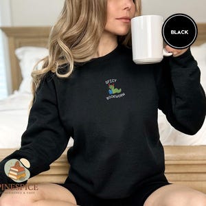 Puede incluir: Sudadera negra con la frase bordada "SPICY BOOKWORM" y un gusano de dibujos animados leyendo un libro. Se sostiene una taza blanca. El logotipo de la marca es visible en la manga izquierda.