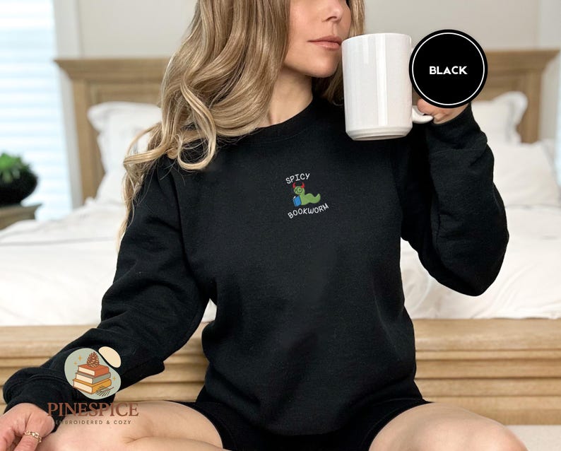 Puede incluir: Sudadera negra con la frase bordada "SPICY BOOKWORM" y un gusano de dibujos animados leyendo un libro. Se sostiene una taza blanca. El logotipo de la marca es visible en la manga izquierda.