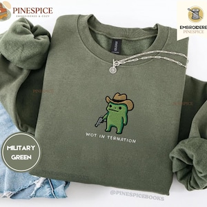 Könnte beinhalten: Ein militärgrüner Rundhals-Pullover mit einer Stickerei eines grünen Frosches, der einen braunen Cowboyhut trägt und eine kleine silberne Pistole hält. Unter dem Frosch steht der Text WOT IN TERNATION in einer einfachen schwarzen Schrift.