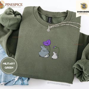 May include: A military green crewneck sweatshirt featuring an embroidered graphic of a large gray creature holding a purple flower and a smaller white creature holding a purple flower bud. The sweatshirt has long sleeves and a ribbed collar.