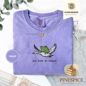 Könnte beinhalten: Violettes Comfort Colors T-Shirt mit einem gestickten Design einer grünen Frosch, der auf einer weißen Gans reitet. Der Text "WE RIDE AT DAWN" steht darunter. Eine goldene Halskette liegt auf dem Shirt.