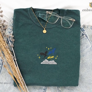May include: A dark green t-shirt with an embroidered design of three dragons flying over an open book. The design is in shades of blue, yellow, and black. The t-shirt is laid flat on a white surface.
